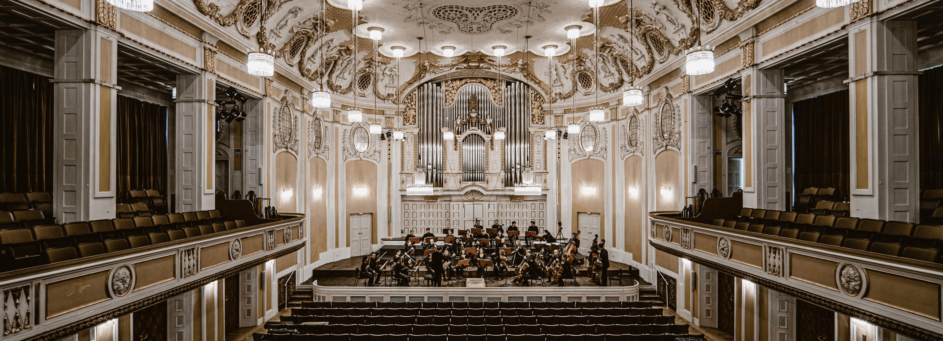 Prunkvoller Konzertsaal mit klassischer Architektur, mit einer großen Orgel auf der Bühne, Kristallleuchtern und gepolsterten Sitzplätzen, während ein Live-Orchester probt.